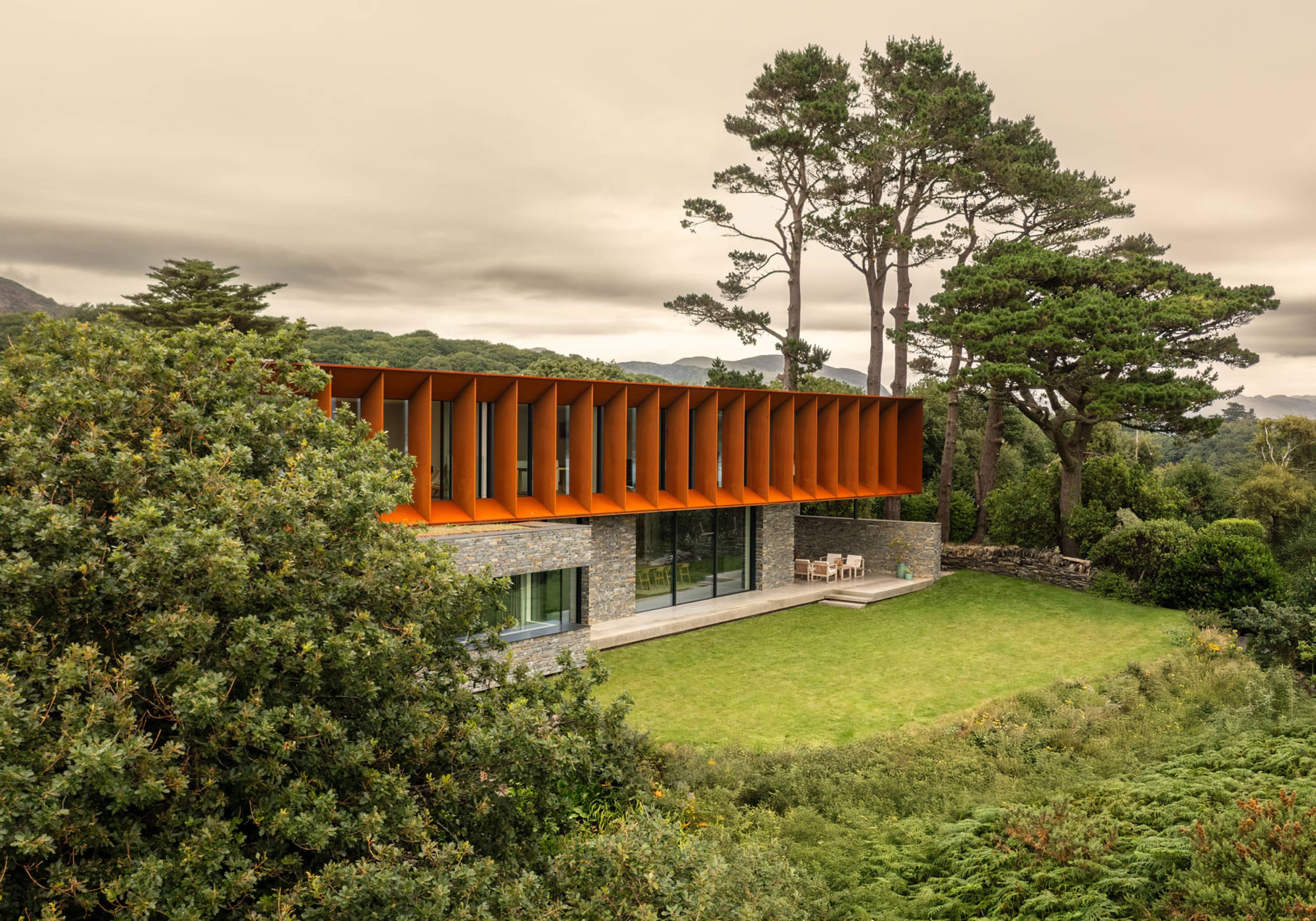 Porthmadog House: a weathering steel upper floor with vertical fins, cantilevered over a slate-clad ground floor, hills behind